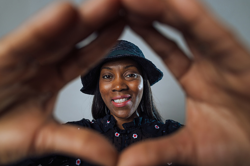 Close-up studio portrait of a mid-female adult holding up her hands with a focus on the woman. She has a cheerful expression on her face. She is wearing a black blouse and a black hat.