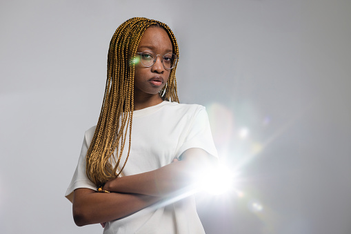 Waist-up studio portrait of a young female adult looking into the camera with a negative expression and her arms crossed. A light from behind her creates a flare into the lens.