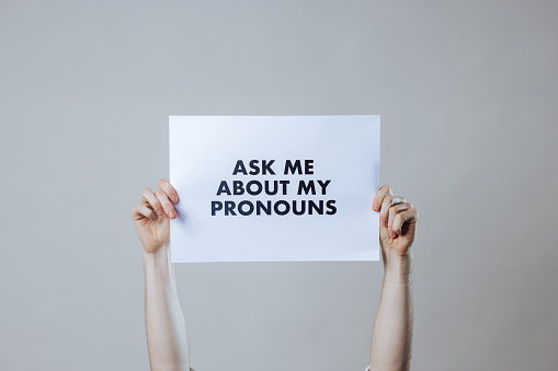 A cropped shot of an unrecognisable person's arms holding a white sign displaying the words ask me about my pronouns