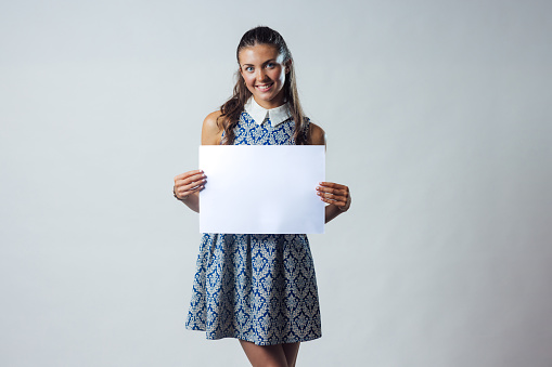 Waist-up studio portrait of a teenage model looking into the camera with a cheerful expression, showing her teeth. She is holding a white sign displaying nothing on it.