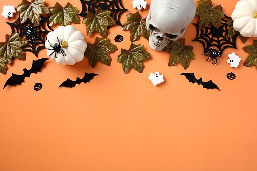 Halloween holiday background with skull, pumpkins, bats, maple leaves, spiders, ghosts on pastel orange table. Flat lay, top view.