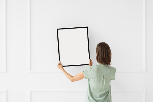 Young woman hanging an artwork, blank empty frame mockup