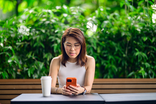 Portrait of a smiling, beautiful Asian businesswoman looking at smartphone, enjoying coffee at a cafe outdoors with a natural background. Copy space