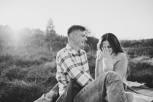 Couple sitting on blanket, hugging in grass in field at sunset. Happy young woman and man spending time together in nature. Concept family holiday. Female embrace male on picnic. Black and white photo