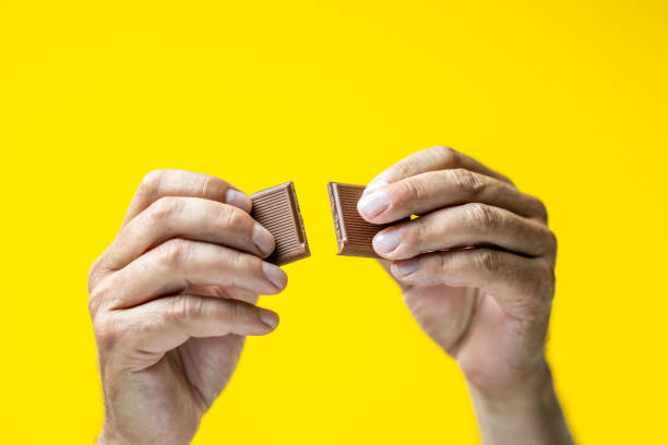 Human hands breaking chocolate Close-up of man's hands breaking piece of chocolate against yellow background. man hand holding candy bar stock pictures, royalty-free photos & images