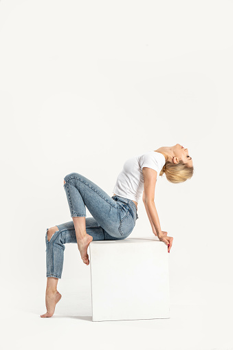 Full length photo of caucasian blonde young woman in casual outfit, white tshirt and jeans, posing in studio. A lot of copy space. Side view. Girl sitting on the cube.