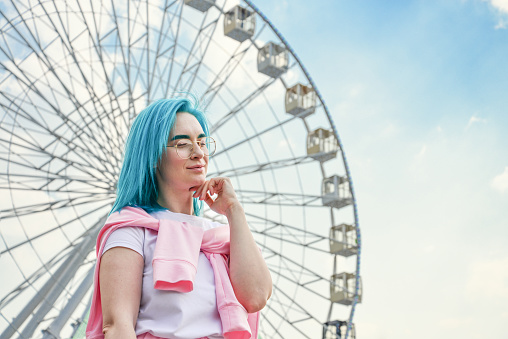 Portrait of young woman with blue hair and stylish sunglasses with cloudy sky and ferris wheel in the background