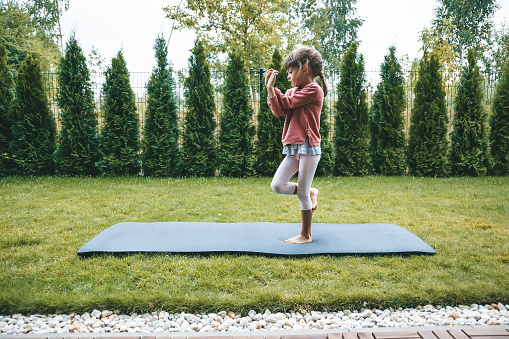 Cute little girl 5-6 years old standing in the Eagle position or Garudasana while practicing yoga outdoors. Yoga for children concept.