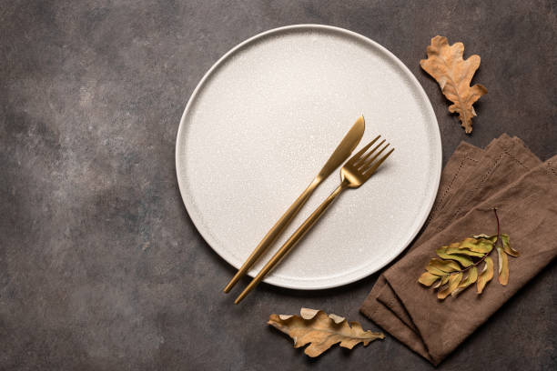 istock Autumn minimal table setting on brown background. Empty beige plate, golden cutlery, linen napkin and dry autumn leaves. Top view, flat lay. 1673854678