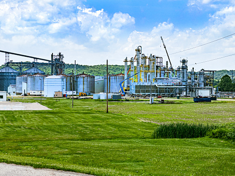 An Ethanol refinery plant is surrounded by a green field and blue sky. The image features silos and buildings surrounded by lush greenery. There are cranes and other construction equipment in the image.