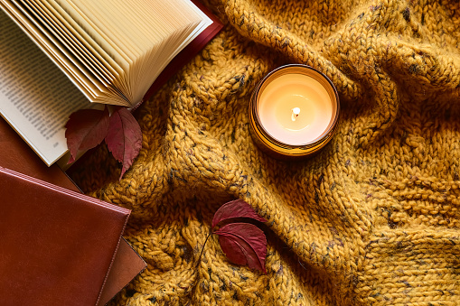 Reading in fall season. Burning candle and open book on knitted plaid with autumn leaves as concept of literature, learning and education in cozy fall mood.