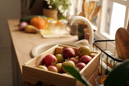 Focus on wooden box with red ripe apples standing in front of camera next to basket with rolled clean towels against sink on kitchen counter