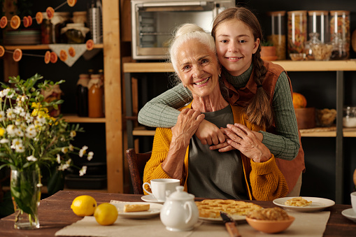 Cute cheerful girl with pigtails giving hug to her grandmother sitting by table with homemade apple pie and tea while both looking at camera