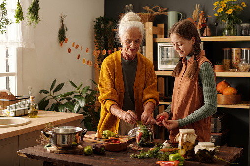 Cute girl with fresh tomatoes helping her grandmother with preparing pickles for winter while looking at senior woman putting vegetables into jar