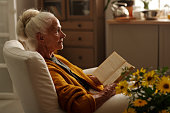 Serious grandmother with open book waiting for someone while sitting in armchair