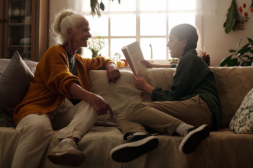 Happy elderly woman in casualwear sitting on couch in front of her granddaughter reading story or novel aloud on autumn weekend