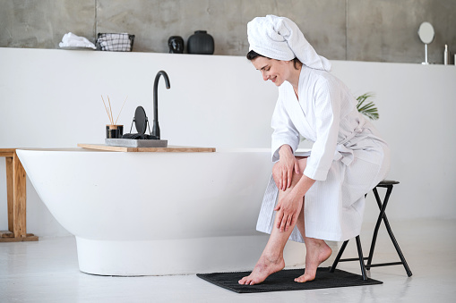 Young woman in white bathrobe and towel on head sitting and touching smooth legs after shave in bathroom. Female doing depilation and applying moisturizer cream for soft skin. Bodycare concept.