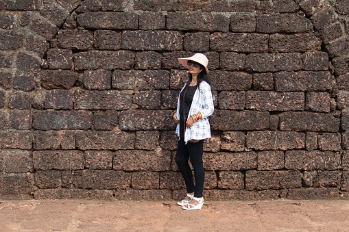 Stylish woman in summer hat and sunglasses posing in front of ancient stone wall. Beautiful female traveler enjoying her vacation at historic site