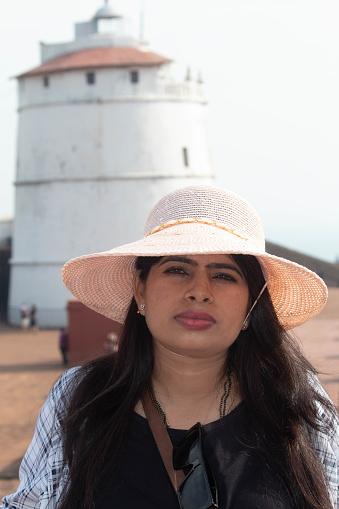 Portrait of stylish female traveler in sun hat. She is looking at camera while standing in front of ancient lighthouse