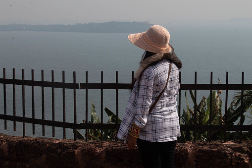 Rear view of young woman admiring seascape from fenced terrace. Stylish female traveler in summer hat looking at beautiful sea during vacation