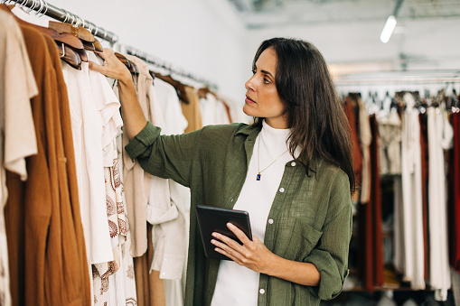 Clothing store owner conducting a stock take in her shop with the help of a trusty tablet. Focused business woman checking and counting items to ensure her inventory is accurate and up-to-date