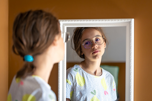 Sleepy teen girl in glasses looks at herself in the mirror in the morning, the concept of self-acceptance.