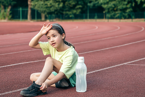 A cute sporty little girl with bottle of water sitting on running track at the stadium, copy space.