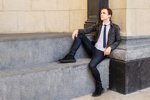 fashionable attractive young man sits against the backdrop of a stone wall, looks thoughtfully to the side with a serious expression on his face