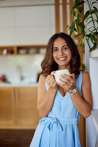 A brunette woman posing for the camera, holding a cup of coffee, being at home.