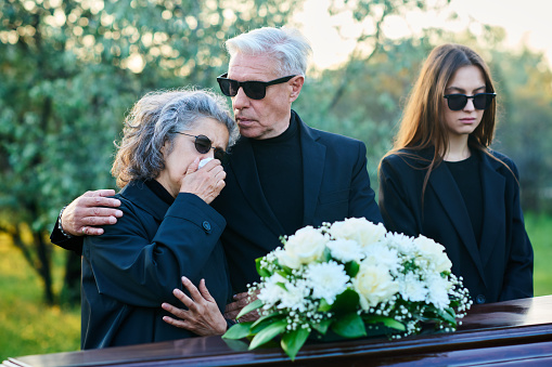 Mature man in black attire and sunglasses consoling his wife lamenting dead relative or family member at funeral while standing in front of coffin