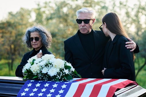 Mature man in mourning attire embracing his grieving daughter while standing by coffin with white flowers on top of closed lid at funeral