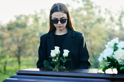 Young widow with bunch of fresh white roses standing by coffin with closed lid and flowers on its top while grieving about her dead husband