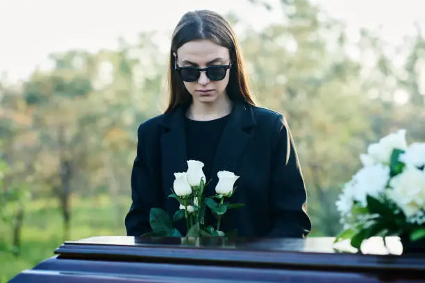 Photo of Young widow with bunch of fresh white roses standing by coffin with closed lid