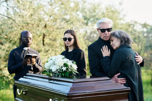 Photo of Mature man supporting his inconsolable wife while both grieving at funeral