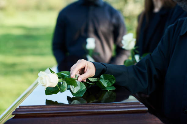 Hand of mature woman in mourning attire putting white rose on coffin lid stock photo