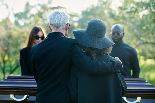 Rear view of mature grey haired man embracing his wife in black hat while both standing in front of coffin and grieving people at funeral