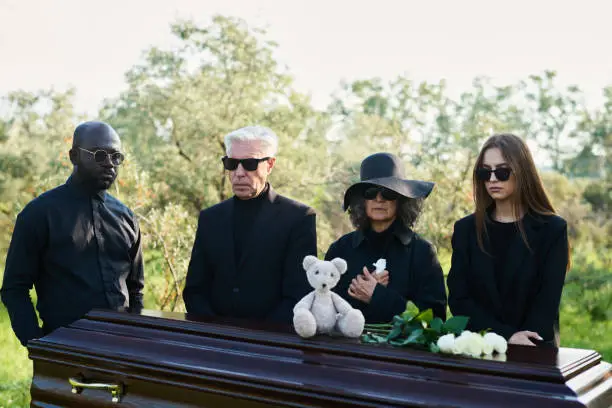 Photo of Group of intercultural people gathered by coffin with teddybear and roses on top