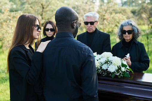 Back view of young woman in mourning attire supporting her husband at funeral while both standing in front of coffin with bunch of white flowers