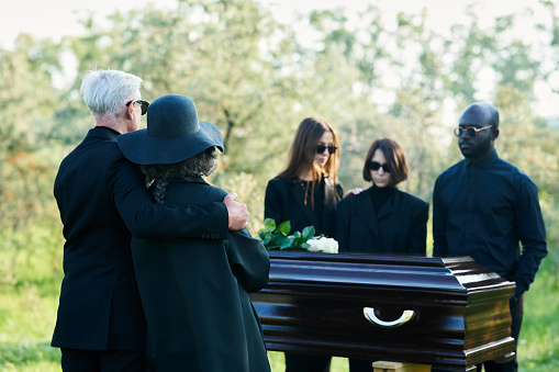 Focus on mature mourning couple wearing black attire standing in front of coffin and group of young intercultural grieving people at funeral