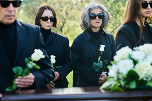 Focus on young and mature women in mourning attire and sunglasses holding white roses while standing in front of coffin at funeral
