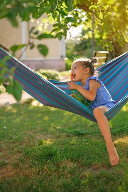 istock A barefoot girl bites off a ripe carrot, laughing merrily while sitting in a hammock under a tree in the yard 1674058065