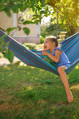 A barefoot girl bites off a ripe carrot, laughing merrily while sitting in a hammock under a tree in the yard