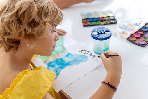 Teacher At Art School Helping Children in Art Class with painting. Woman teaches child to create and work with aquarelle paint. Group of primary school kids learning how to draw and color help of their educator.