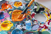 Artist's hand picks up acrylic or oil paint from palette on brush. Closeup paintbrush picking orange color from an artist palette. Colorful image from an artist’s studio or a school showing creative education.