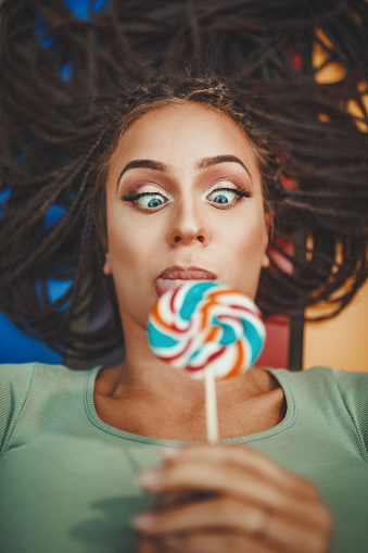 Cheerful young Caucasian woman with beautiful long afro braided hair is licking the lollipop, having fun and looking on it.