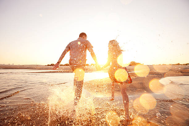 una felice coppia attraversa onde sulla soleggiata spiaggia - heterosexual couple couple sensuality young couple foto e immagini stock