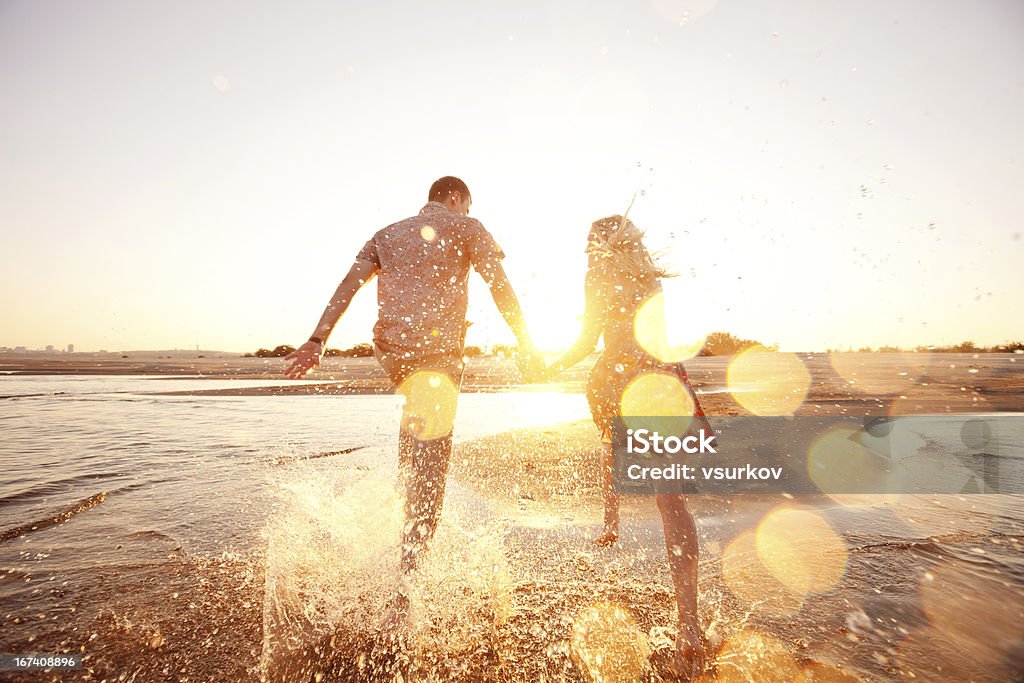 A happy couple runs through waves on sunlit beach happy couple running on the beach Couple - Relationship Stock Photo
