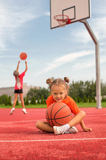 Charming female child basketball player holding game ball and smiling while standing against light orange background