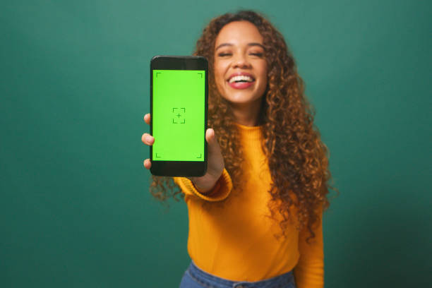 Happy young woman holds up device chroma key screen, green studio background stock photo