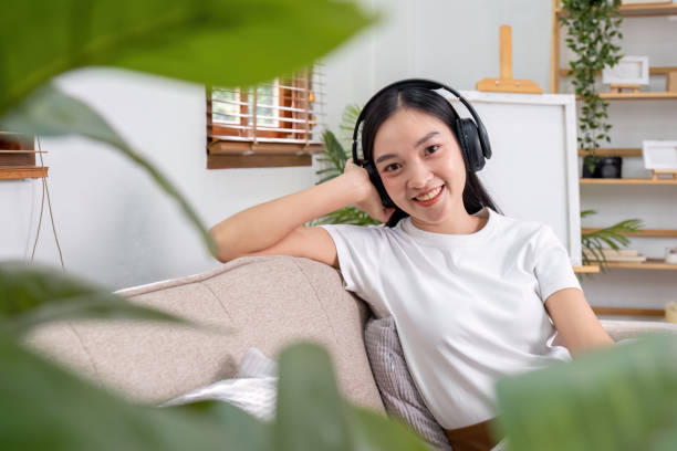 jovem mulher ouvindo música enquanto sentada em um sofá de casa usando fones de ouvido se sente feliz e relaxada passando o tempo livre em casa. - room service audio - fotografias e filmes do acervo